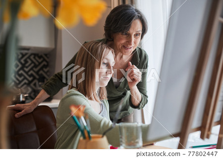 Woman looking with satisfaction at the drawing of her blonde granddaughter Woman looking with satisfaction at the drawing of her blonde granddaughter 77802447