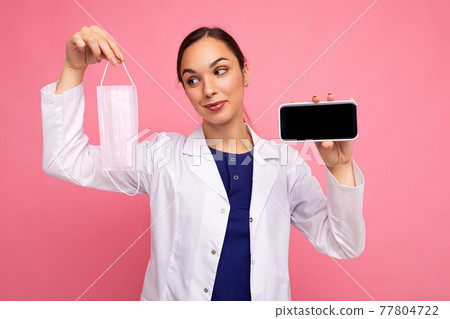pretty female doctor wearing white coat holding mask showing modern mobile phone isolated over pink 77804722