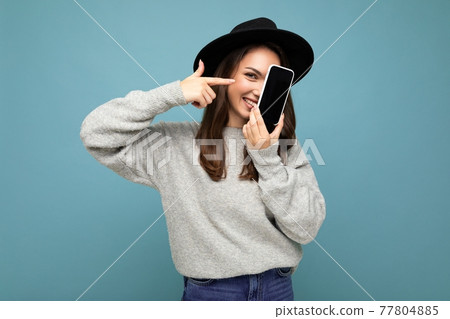 Beautiful positive woman wearing black hat and grey sweater holding mobilephone showing smartphone 77804885