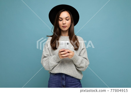 Beautiful young brunette woman wearing black hat and grey sweater holding smartphone looking down at 77804915