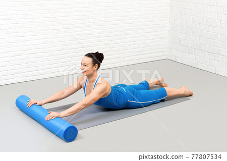 Back extention drill with foam roller. Adult smiling caucasian woman in blue sportswear lies on her stomach and does pilates workout in loft fitness studio, indoor. Back extention drill with foam roller. Adult smiling caucasian woman in blue sportswear lies on her stomach and does pilates workout in loft fitness studio, indoor. 77807534