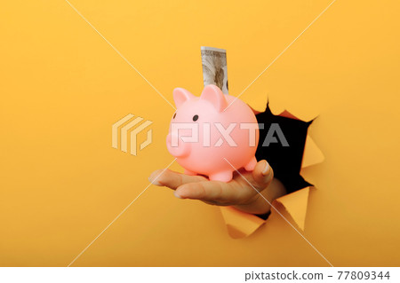 Hand with a piggy bank and dollar banknote through a yellow paper hole. Investment and savings Hand with a piggy bank and dollar banknote through a yellow paper hole. Investment and savings 77809344
