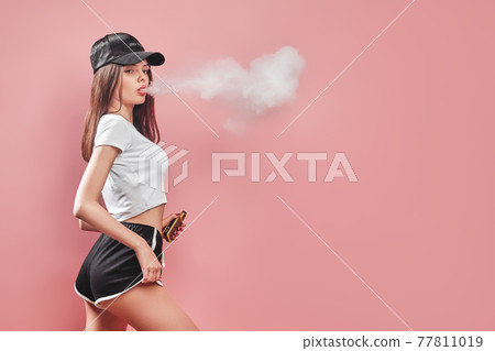 Seductive glamour young woman standing and vaping on pink background 77811019