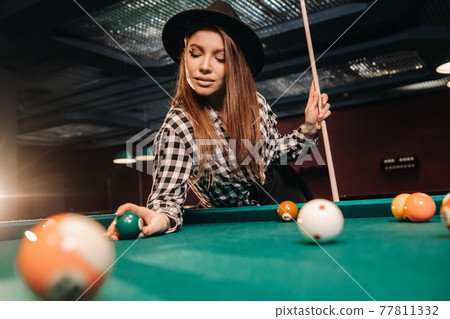 A girl in a hat in a billiard club with a cue and balls in her hands.Playing pool 77811332