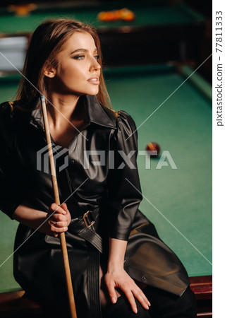 A girl with a cue in her hands sits on a table in a billiard club.Russian billiards 77811333