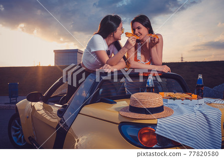 Two young females eating pizza, smiling, posing in yellow car with french fries, hat and soda in glass bottle on its trunk. Fast food. Mock up 77820580