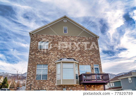 Gabled home with bay window balcony and stone brick wall against cloudy blue sky Gabled home with bay window balcony and stone brick wall against cloudy blue sky 77820789