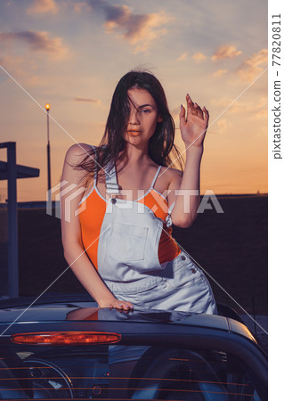 Sexy young female in white overall and orange t-shirt is posing outdoors standing in car cabriolet. Summer evening, sunset. Close up Sexy young female in white overall and orange t-shirt is posing outdoors standing in car cabriolet. Summer evening, sunset. Close up 77820811