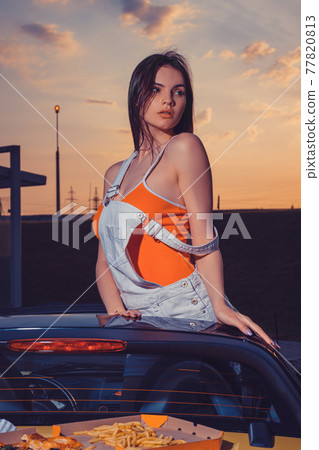 Sexy woman in white overall and orange t-shirt is posing standing in yellow car cabrio with french fries and pizza on its trunk. Fast food. Close up Sexy woman in white overall and orange t-shirt is posing standing in yellow car cabrio with french fries and pizza on its trunk. Fast food. Close up 77820813