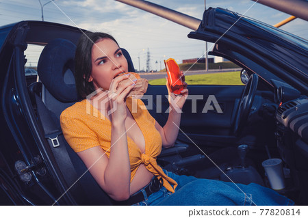 Female in blue jeans and orange top is eating hamburger and holding paper package with french fries while sitting in yellow car. Fast food. Copy space Female in blue jeans and orange top is eating hamburger and holding paper package with french fries while sitting in yellow car. Fast food. Copy space 77820814