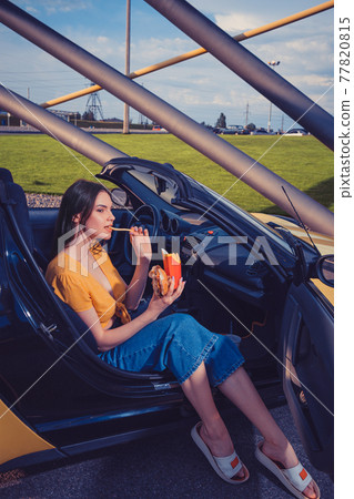 Woman in blue jeans and orange top is eating french fries from paper package and holding hamburger while sitting in yellow car cabrio Copy space Woman in blue jeans and orange top is eating french fries from paper package and holding hamburger while sitting in yellow car cabrio Copy space 77820815