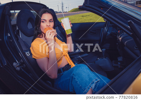 Model in blue jeans and orange top is eating hamburger and holding beverage in paper cup while sitting in yellow car cabriolet. Fast food. Copy space Model in blue jeans and orange top is eating hamburger and holding beverage in paper cup while sitting in yellow car cabriolet. Fast food. Copy space 77820816
