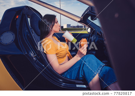 Lady in blue jeans and orange top is drinking beverage from paper cup, holding hamburger while sitting in yellow car cabrio. Fast food. Mock up Lady in blue jeans and orange top is drinking beverage from paper cup, holding hamburger while sitting in yellow car cabrio. Fast food. Mock up 77820817