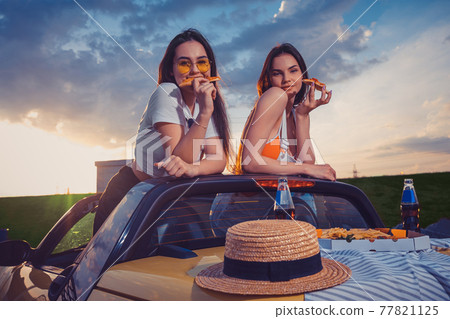 Charming girls eating pizza, posing in yellow car roadster with french fries, hat and soda in glass bottles on trunk. Fast food. Mock up 77821125