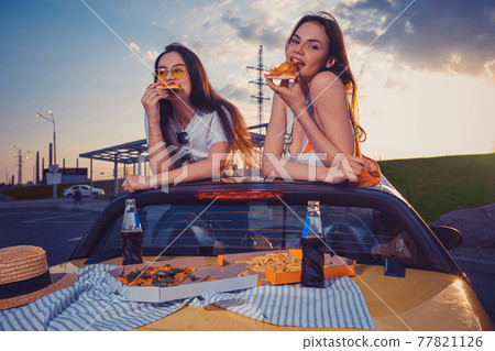Charming females are eating pizza while posing in yellow car cabriolet with french fries and soda in glass bottles on trunk. Fast food. Close up 77821126