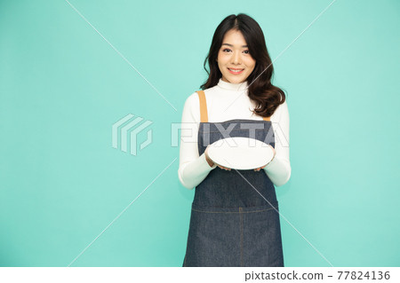 Young Asian woman in apron standing and holding empty white plate or dish isolated on green background 77824136