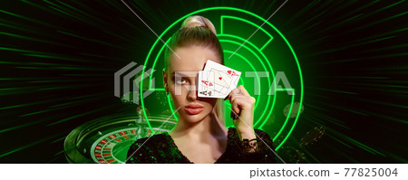 Woman in black dress. Covered eye by two aces, posing on black background with neon lights, roulette and falling chips. Poker, casino. Close-up 77825004