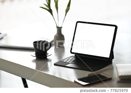 A computer tablet with a white blank screen is putting on a working desk surrounded by various equipment. A computer tablet with a white blank screen is putting on a working desk surrounded by various equipment. 77825970