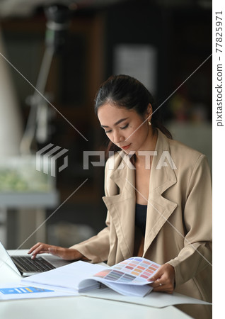 Designer woman is using a computer laptop while sitting and selecting from a color guide at the working desk. Designer woman is using a computer laptop while sitting and selecting from a color guide at the working desk. 77825971