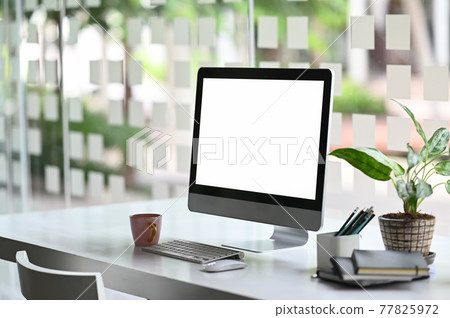 A computer monitor with a white blank screen is putting on a working desk surrounded by various equipment. A computer monitor with a white blank screen is putting on a working desk surrounded by various equipment. 77825972