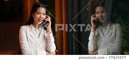 A businesswoman is talking on a mobile phone while standing over an office glass wall. A businesswoman is talking on a mobile phone while standing over an office glass wall. 77825977