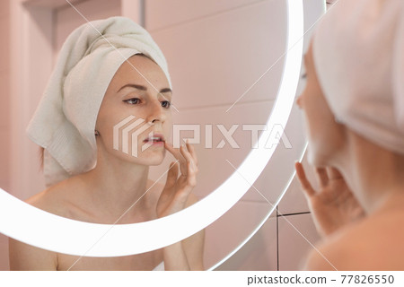 Woman standing near mirror in the bathroom and applying moisturizer her face Woman standing near mirror in the bathroom and applying moisturizer her face 77826550