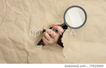 a woman's hand holds a glass magnifier, part of the body sticks out of a hole in brown paper a woman's hand holds a glass magnifier, part of the body sticks out of a hole in brown paper 77829009