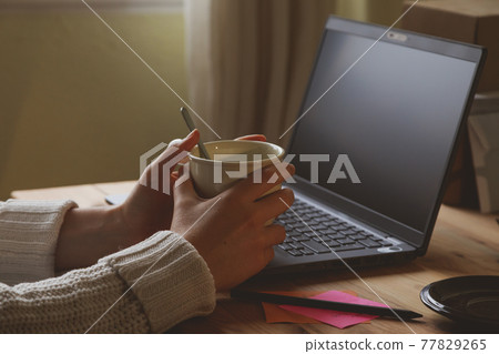 Photograph of the desk and the hands of a young woman, drinking a hot coffee or infusion, early in the morning, while teleworking at home, in Madrid, Spain. Photograph of the desk and the hands of a young woman, drinking a hot coffee or infusion, early in the morning, while teleworking at home, in Madrid, Spain. 77829265
