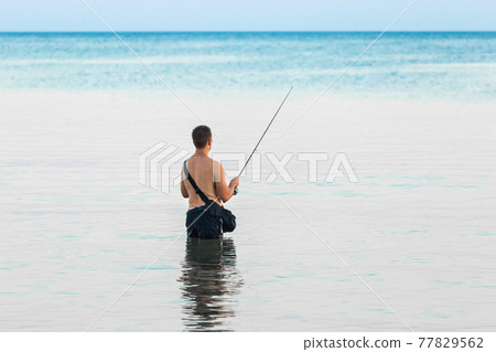 A young guy without a shirt stands knee-deep in the water of the sea in the evening and catches fish with a line A young guy without a shirt stands knee-deep in the water of the sea in the evening and catches fish with a line 77829562
