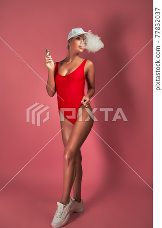 Seductive glamour young woman in red swimsuit standing and vaping on pink background 77832037