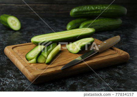 Board with ripe cucumber and knife on black smokey table 77834157