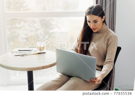 Beautiful young woman working on laptop computer while sitting at the living room, drinking coffee. Home office during Coronavirus or Covid-19 quarantine. Communicates on internet. 77835536