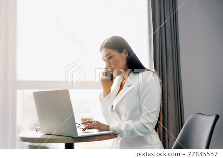 Beautiful woman talking on phone while working on laptop computer sitting at home managing her business via home office during Coronavirus or Covid-19 quarantine Beautiful woman talking on phone while working on laptop computer sitting at home managing her business via home office during Coronavirus or Covid-19 quarantine 77835537
