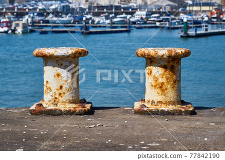 Two pillar bollards for mooring boats with the harbour in the background Two pillar bollards for mooring boats with the harbour in the background 77842190