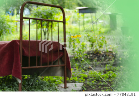Rest in the village. Blurred background. Defocused An iron bed covered with a blanket in a garden. Cozy relaxation in nature near the house 77848191