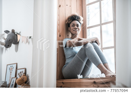 Depressed young dark skinned woman sitting on windowsill 77857094