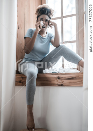 Crying woman sitting at home on windowsill 77857096