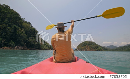 Man with thin plait hairstyle rows pink plastic kayak putting up paddle on sea against hills with wild jungles and blue sky. 77858705