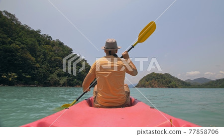 Man with sunglasses and hat rows pink plastic canoe along sea against green hilly islands with wild jungles. Traveling to tropical countries. 77858706