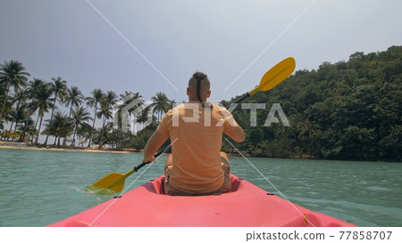 Man with sunglasses and hat rows pink plastic canoe along sea against green hilly islands with wild jungles. Traveling to tropical countries. Man with sunglasses and hat rows pink plastic canoe along sea against green hilly islands with wild jungles. Traveling to tropical countries. 77858707