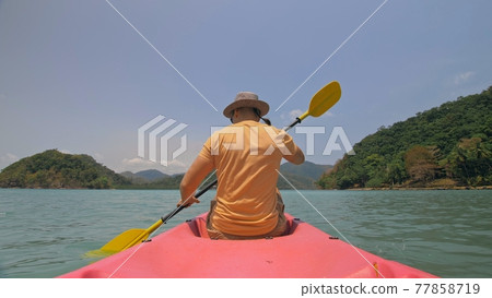 Man with sunglasses and hat rows pink plastic canoe along sea against green hilly islands with wild jungles. Traveling to tropical countries. 77858719
