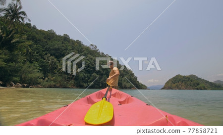 Man with sunglasses and hat rows pink plastic canoe along sea against green hilly islands with wild jungles. Traveling to tropical countries. 77858721