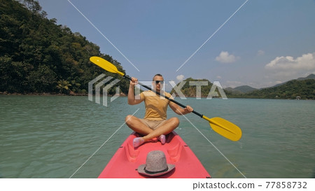 Man with sunglasses and hat rows pink plastic canoe along sea against green hilly islands with wild jungles. Traveling to tropical countries. 77858732