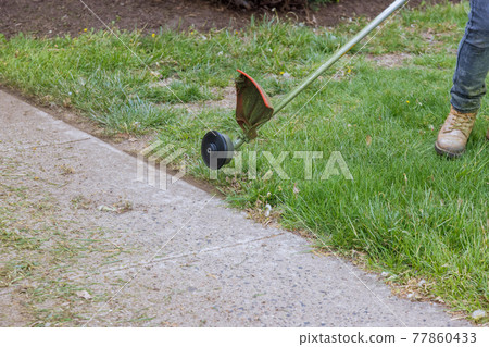 A lawn mower mows fresh, green grass on the lawn near, a municipal worker with lawn mower in his hand. 77860433