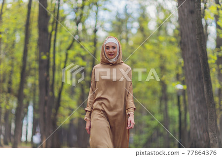 Islamic beautiful woman in a Muslim dress standing on a summer park street background forest autumn trees.world hijab day. 77864376