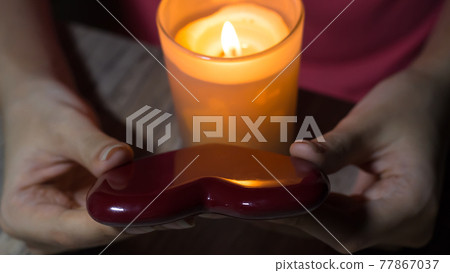 Red heart in the hands of a girl with a burning candle. A fortune teller holds a heart in her hands and guesses at future love with a lighted candle. Christmas fortune telling and fortune telling. 77867037