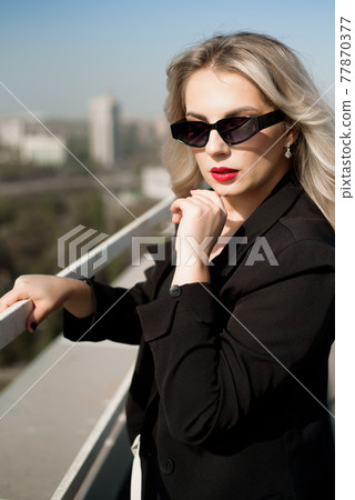 Elegant woman in a black jacket and sunglasses at rooftop bar terrace looking at city. 77870377