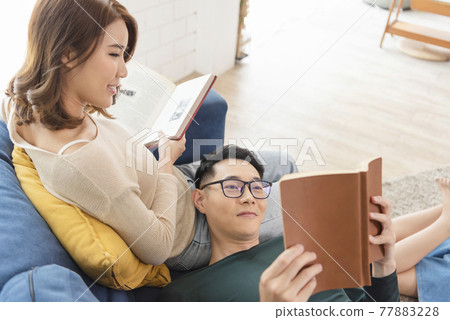 Happy asian couple is spending weekend together on couch indoors at home, relaxing and enjoying reading book. 77883228