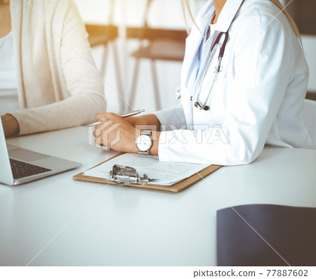 Unknown woman-doctor communicating with female patient in sunny clinic. Physician checks medical history record and exam results, close-up Unknown woman-doctor communicating with female patient in sunny clinic. Physician checks medical history record and exam results, close-up 77887602