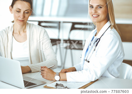 Woman-doctor sitting and communicating with her female patient in clinic. Blonde physician happy to help. Medicine concept Woman-doctor sitting and communicating with her female patient in clinic. Blonde physician happy to help. Medicine concept 77887603
